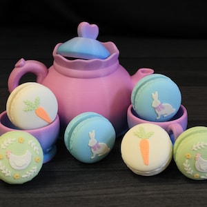 May include: A whimsical Easter-themed arrangement featuring a pink teapot with a blue lid, surrounded by pastel-colored macarons. The macarons are decorated with carrots, bunnies, and chickens, presented in small purple teacups.