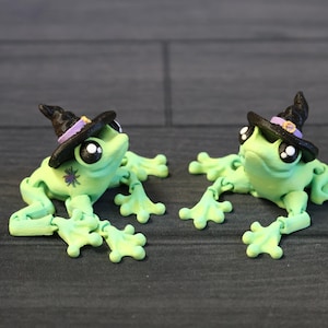 May include: Two green frog figurines wearing black witch hats with purple trim. The frogs have large black eyes and are posed with their legs outstretched.