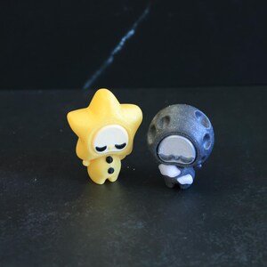 May include: Two small figurines: one yellow star-shaped character with a smooth face and two black buttons on its body, and one dark gray, textured, cratered moon-shaped character with a white face and small white arms held across its chest.