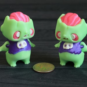 May include: Two green zombie figurines with pink brains, purple vests with skull emblems, and white eyes. A coin is placed between the figurines. The figurines have small arms and are standing.