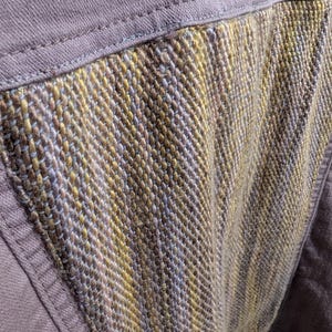 May include: Close-up of a section of a mauve-colored garment with a textured, woven panel. The panel features a mix of gold, brown, and blue threads, creating a unique pattern. The garment's seams are visible, adding to the handcrafted aesthetic.