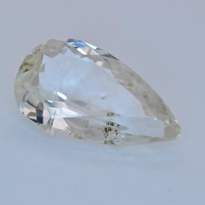 May include: A clear, pear-shaped gemstone with multiple facets. The gem has a slight yellow tint and internal inclusions. The cut of the stone creates a play of light and shadow, enhancing its brilliance. The gemstone is set against a white background.