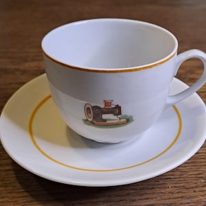 Könnte beinhalten: Eine weiße Teetasse und Untertasse mit goldfarbenem Rand. Die Teetasse zeigt ein Bild einer alten Nähmaschine.