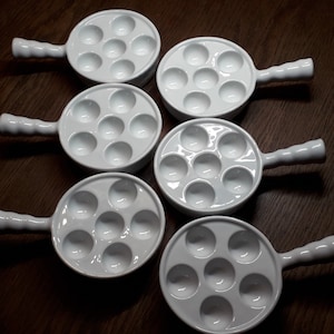 Könnte beinhalten: Set aus sieben weißen Keramik-Ramekins mit Griffen. Jedes Ramekin hat sieben einzelne Vertiefungen zum Kochen oder Servieren kleiner Portionen von Speisen.