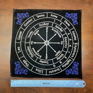 May include: A black velvet Ouija board with a white circle in the center and a white star. The circle is divided into 12 sections, each with a zodiac sign and a day of the week. The board has blue embroidered corners.