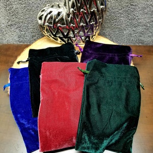 May include: Five velvet drawstring pouches in different colors: blue, black, red, purple, and green. The pouches are arranged on a wooden surface.