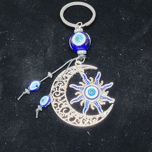 May include: Silver key chain with a crescent moon and sun design. The moon and sun have blue and white evil eye charms. The key chain also has a blue and white evil eye bead.