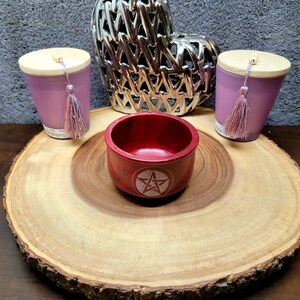 May include: A red wooden bowl with a white pentagram design sits on a wooden slice. Two purple candles with wooden lids and tassels are on either side of the bowl. A silver heart-shaped decorative object is behind the candles.