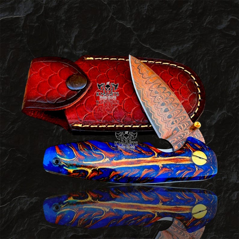May include: A folding pocket knife with a Damascus steel blade and a blue and copper resin handle, accompanied by a red leather sheath featuring a scale pattern and the text KN.GHTS. The knife has a gold-colored pivot screw.