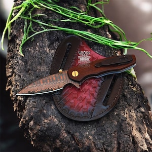 May include: A folding pocket knife with a patterned Damascus steel blade, a dark wood handle, and a gold-colored pivot screw. The knife features a black pocket clip and rests on a round, red and brown embossed leather sheath with the text KNIGHTS.