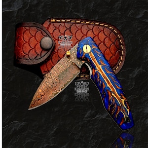May include: A folding pocket knife with a Damascus steel blade featuring a wavy pattern and a blue and gold resin handle. The knife is positioned in front of a brown leather sheath with a scale-like embossed texture and a metal snap button.