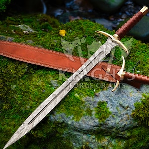 May include: An intricately patterned sword with gold detailing and a brown leather handle, alongside its brown leather sheath. The sword is displayed on a moss-covered rock. The image has a natural, outdoor setting.