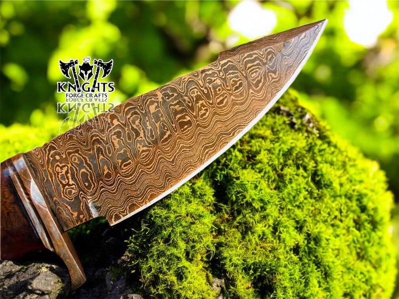 Puede incluir: Primer plano de una hoja de cuchillo de acero de Damasco con un patr&oacute;n &uacute;nico. La hoja es marr&oacute;n y plateada, con un mango de madera. El cuchillo descansa sobre musgo verde. El logotipo "KNIGHTS FORGE CRAFTS" es visible.