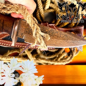 Puede incluir: Un cuchillo artesanal con una hoja estampada y un mango de madera, en una funda de cuero marr&oacute;n. El cuchillo se sujeta con una cuerda y el logotipo "KNIGHTS FORGE CRAFTS" es visible. Flores blancas en primer plano.