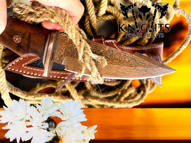Puede incluir: Un cuchillo artesanal con una hoja estampada y un mango de madera, en una funda de cuero marr&oacute;n. El cuchillo se sujeta con una cuerda y el logotipo "KNIGHTS FORGE CRAFTS" es visible. Flores blancas en primer plano.