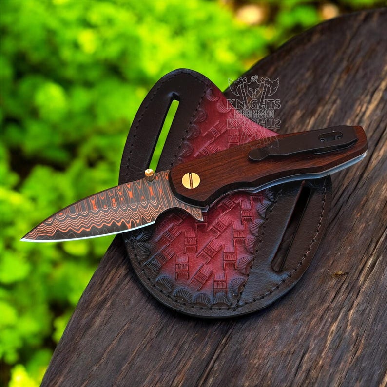 May include: A folding pocket knife with a patterned Damascus steel blade and a dark wood handle featuring a gold-colored screw. The knife rests on a circular, dark brown and red leather sheath with a stamped geometric pattern and a logo.