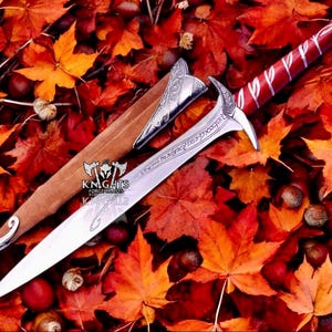 May include: A silver and red sword with a brown sheath, set against a backdrop of autumn leaves. The sword has an ornate handle and blade with engraved text. The sheath features silver detailing. The scene is filled with red and orange leaves.