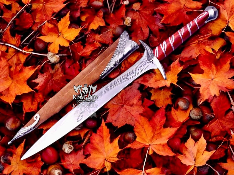 May include: A silver and red sword with a brown sheath, set against a backdrop of autumn leaves. The sword has an ornate handle and blade with engraved text. The sheath features silver detailing. The scene is filled with red and orange leaves.