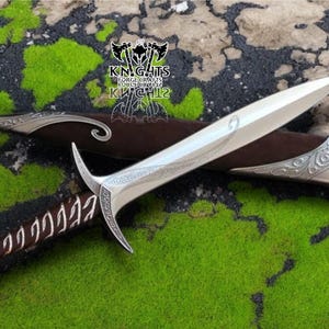 May include: A silver dagger with a curved blade and ornate handle in a brown leather sheath. The dagger has decorative engravings and a curved guard. The sheath has silver accents. The background is green and brown.