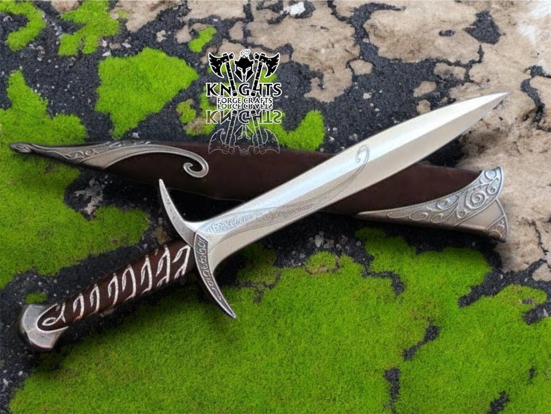 May include: A silver dagger with a curved blade and ornate handle in a brown leather sheath. The dagger has decorative engravings and a curved guard. The sheath has silver accents. The background is green and brown.