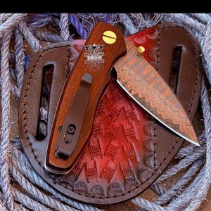 May include: A folding pocket knife with a wood handle featuring a gold screw and a black pocket clip. The blade has a copper-toned Damascus pattern. The handle displays a logo with two axes and the text KNIGHTS KNIVES. It rests on a round leather sheath.