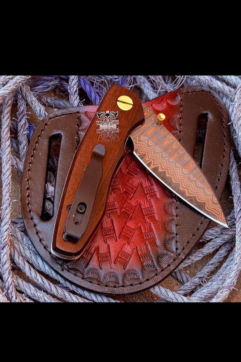 May include: A folding pocket knife with a wood handle featuring a gold screw and a black pocket clip. The blade has a copper-toned Damascus pattern. The handle displays a logo with two axes and the text KNIGHTS KNIVES. It rests on a round leather sheath.