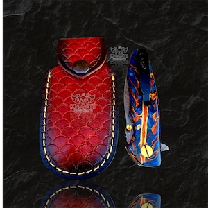 May include: A red leather sheath with a scale pattern and a stamped logo featuring two axes and the text KNIGHTS EDGE, paired with a folding pocket knife featuring a blue and orange marbled handle, a black metal pocket clip, and gold screws.