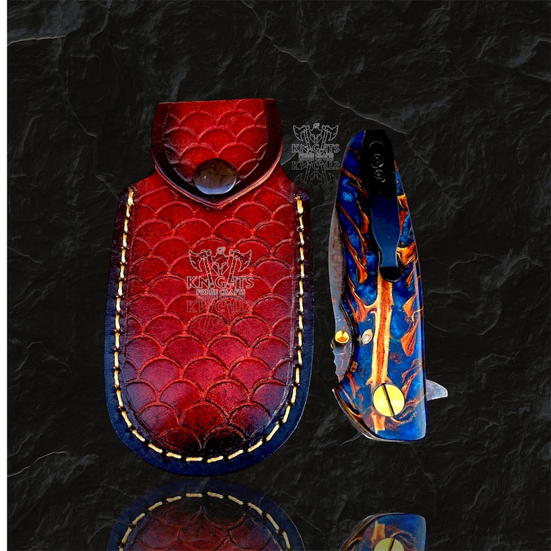 May include: A red leather sheath with a scale pattern and a stamped logo featuring two axes and the text KNIGHTS EDGE, paired with a folding pocket knife featuring a blue and orange marbled handle, a black metal pocket clip, and gold screws.