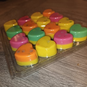 Puede incluir: Una bandeja de plástico transparente llena de fundidos de cera en forma de corazón de varios colores, incluyendo rosa, verde, amarillo y naranja. Los fundidos tienen un acabado brillante y están dispuestos en un patrón escalonado.