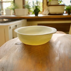 May include: A white ceramic bowl with a yellow glaze sits on a wooden table. The bowl has a rounded shape and a slightly flared rim.