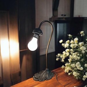 Puede incluir: Una lámpara de mesa de estilo vintage con un brazo curvo y una bombilla blanca. La lámpara tiene un acabado en bronce y una base decorativa.