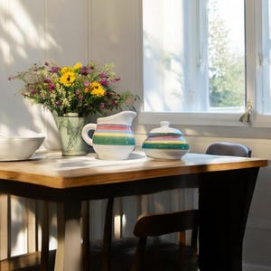 May include: A wooden table with a white pitcher and a sugar bowl with colourful stripes. The table is set with a white bowl and a bouquet of flowers in a vase.