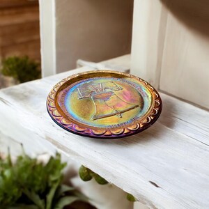 Può includere: Un piatto di vetro iridescente con un'immagine in rilievo della Liberty Bell. Il piatto ha un bordo a festoni ed è decorato con un motivo dorato.