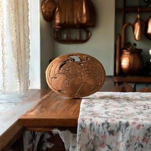 Puede incluir: Un salvamanteles de metal de color cobre con un diseño de fruta, incluyendo manzanas y peras. El salvamanteles está sobre una mesa de madera con un mantel floral.