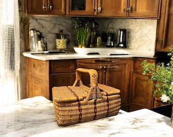 May include: A woven wicker picnic basket with a brown handle and red accents. The basket is sitting on a white marble countertop.