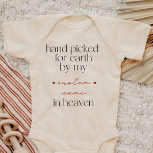 以下が含まれることがあります： 白いベビーロンパースに「hand picked for earth by my custom name in heaven」という赤い文字が書かれています。ロンパースは白と茶色のストライプの毛布の上に置かれています。