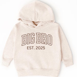 K&ouml;nnte beinhalten: Ein beiger Kapuzenpullover mit dem Aufdruck "BIG BRO EST. 2025" in Braun auf der Vorderseite.