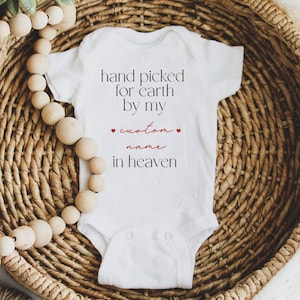 以下が含まれることがあります： 白いベビーロンパースに「hand picked for earth by my [custom name] in heaven」という黒い文字が書かれており、名前の周りに赤いハートが2つあります。