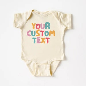May include: A cream-colored baby onesie with the text "YOUR CUSTOM TEXT" in colorful, bubble-like letters.