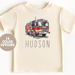 Pode incluir: Uma camiseta bege claro com uma ilustração em aquarela de um caminhão de bombeiros vermelho. O texto "HUDSON" é impresso abaixo do caminhão.