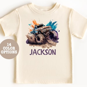 Pode incluir: Uma camiseta bege com um design de aquarela colorido de um caminhão monstro. O nome "JACKSON" está impresso em roxo abaixo do caminhão.