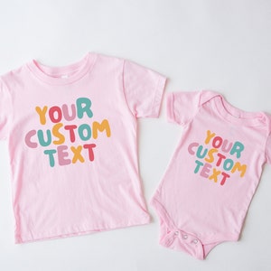 May include: Two pink t-shirts with the text "YOUR CUSTOM TEXT" in colorful, bubble-like letters. The larger shirt has short sleeves and the smaller shirt has a snap closure.