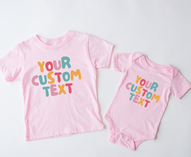 May include: Two pink t-shirts with the text "YOUR CUSTOM TEXT" in colorful, bubble-like letters. The larger shirt has short sleeves and the smaller shirt has a snap closure.
