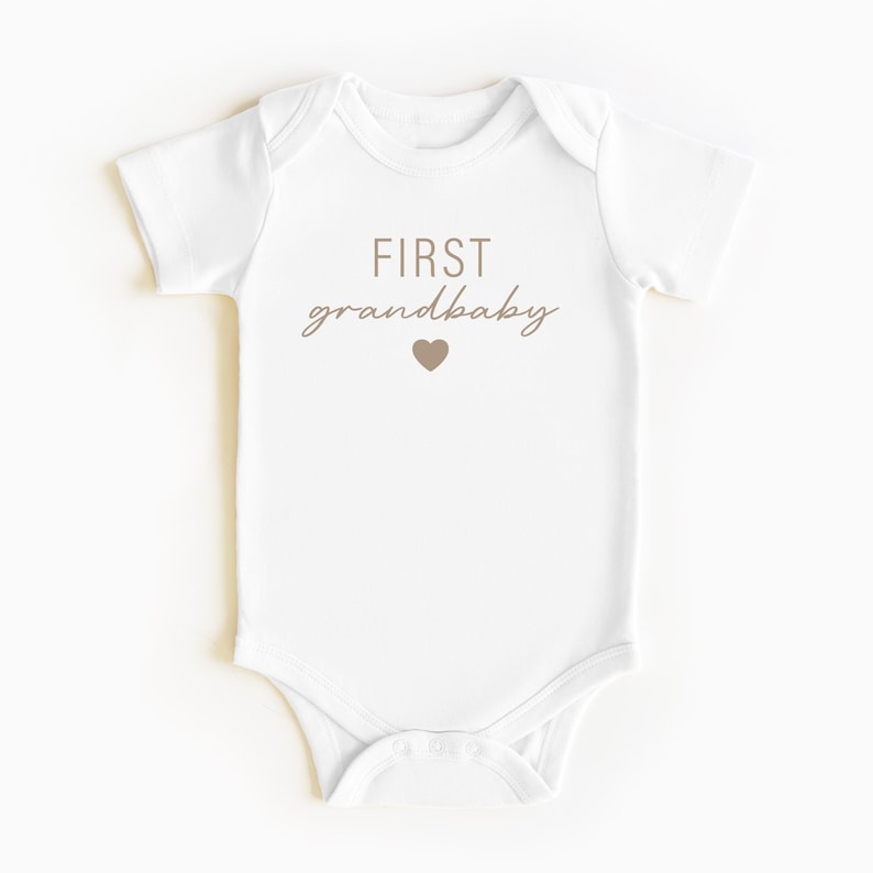 May include: White baby onesie with brown text that reads "FIRST grandbaby" with a small brown heart below the text.