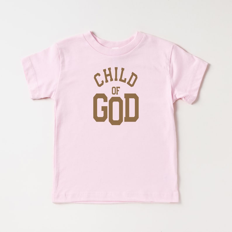 May include: Pink toddler t-shirt with brown text that reads "CHILD OF GOD" in a varsity style font.