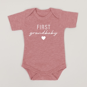 May include: A pink baby onesie with short sleeves and the text "FIRST grandbaby" in white with a white heart.