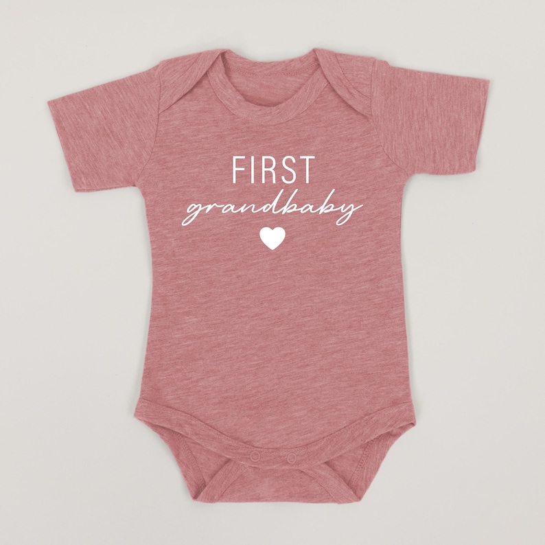 May include: A pink baby onesie with short sleeves and the text "FIRST grandbaby" in white with a white heart.