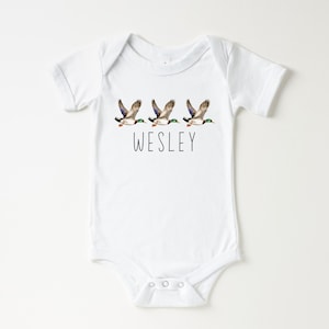 May include: White baby onesie with a design of three flying ducks and the name "WESLEY" printed below.
