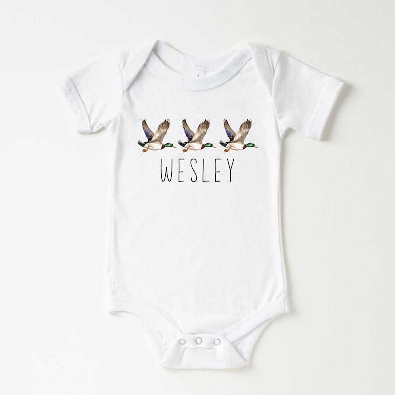 May include: White baby onesie with a design of three flying ducks and the name "WESLEY" printed below.