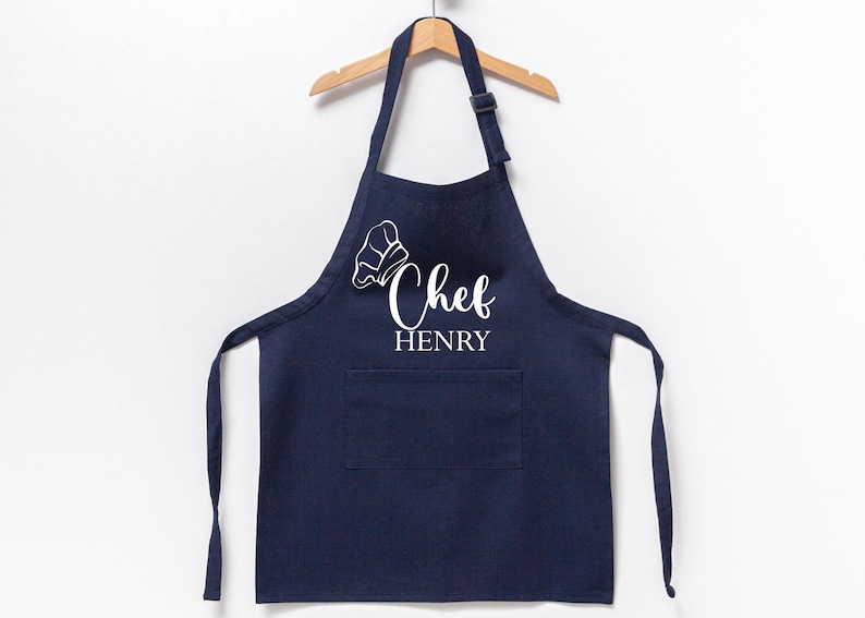 K&ouml;nnte beinhalten: Eine marineblaue Sch&uuml;rze mit einer wei&szlig;en Kochm&uuml;tze und dem Text "Chef HENRY" auf der Vorderseite.